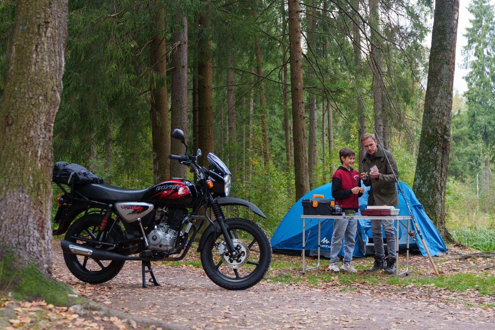 Сын и отец собирают снасти у палатки; мотоцикл Bajaj Boxer слева кадра.
